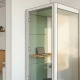 Glazen kantoorpod met houten bureau, gesloten laptop en salie groene akoestische panelen in lichte Nederlandse kantoorruimte.